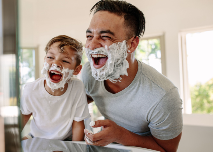 Dad and son with shaving foam on their faces laughing into a mirror