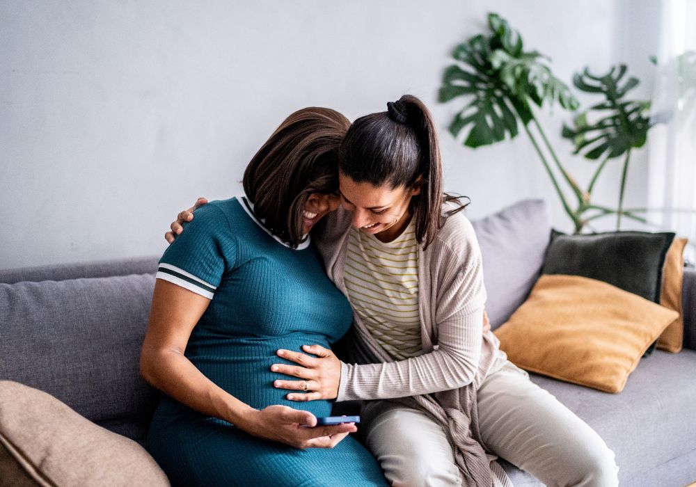Woman hugging pregnant friend who is looking at phone
