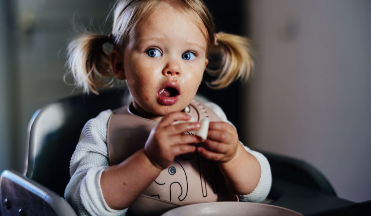 Amazed cute toddler girl with pigtails eating at a table at home