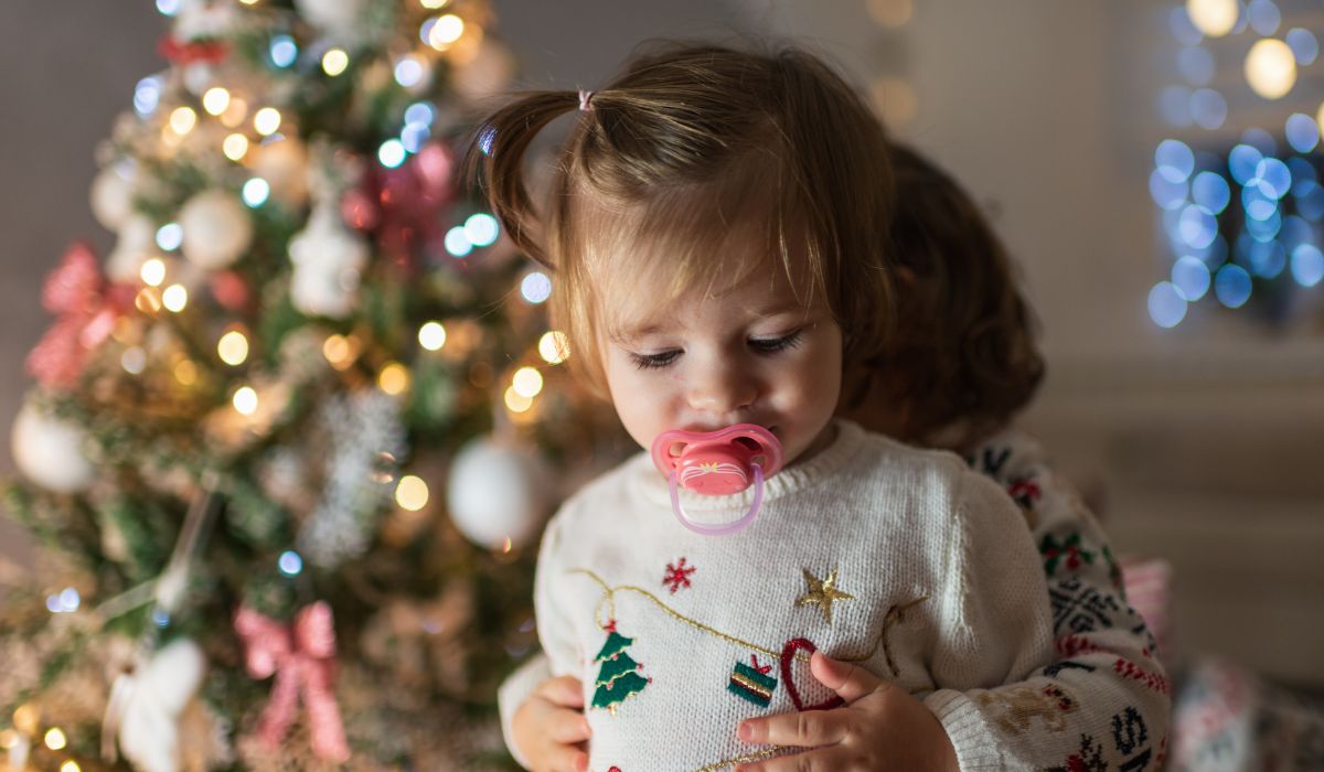Little girl wearing Christmas jumper with soother in mouth being hugged by sibling. Christmas tree in background
