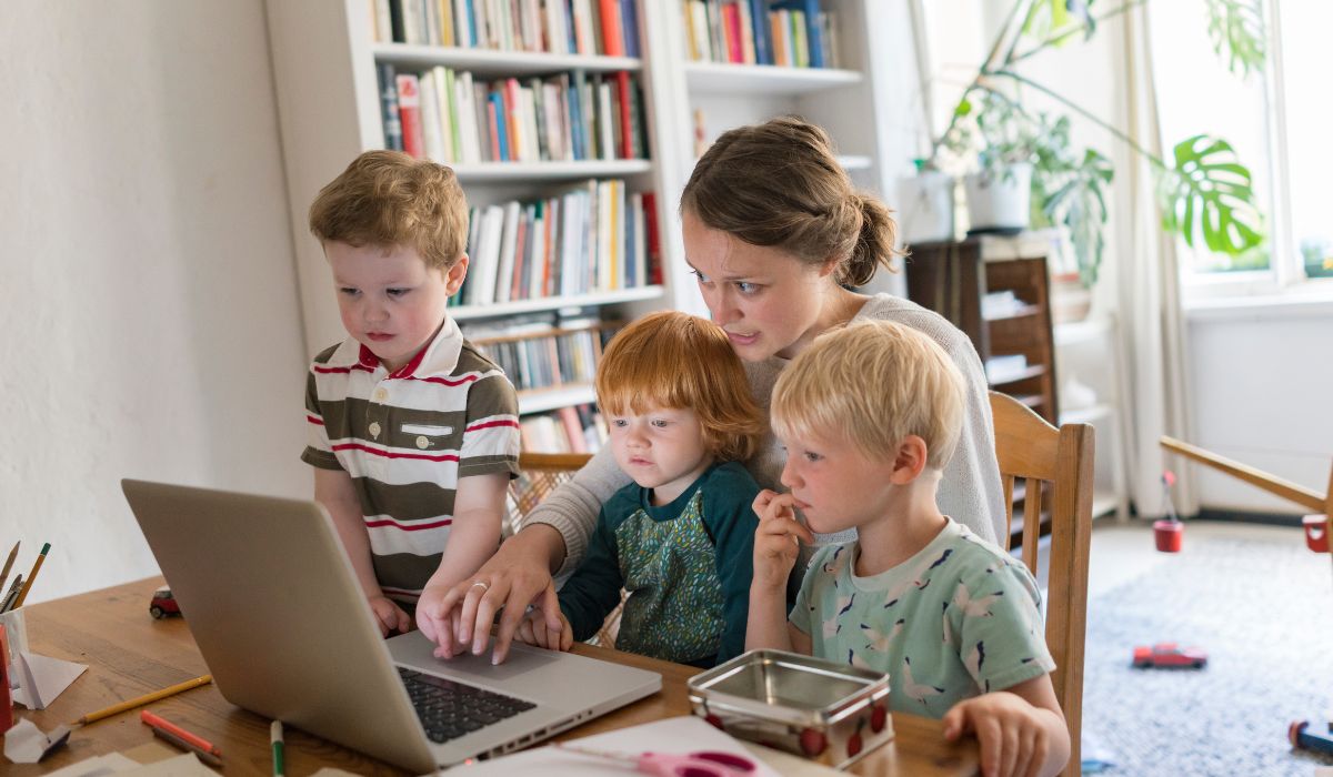Mum on her laptop with a child on her lap, one on her chair and one standing up