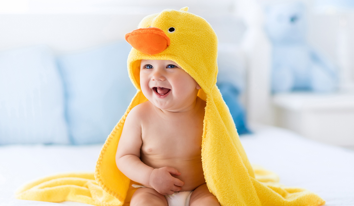 Happy laughing baby wearing yellow hooded duck towel sitting on parents bed