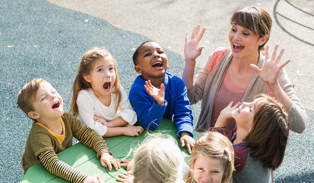 High angle view of a preschool teacher with a multi-ethnic group of children, 4 to 5 years, laughing and having fun. They are sitting at a green table outdoors, mouths open, laughing and shouting.