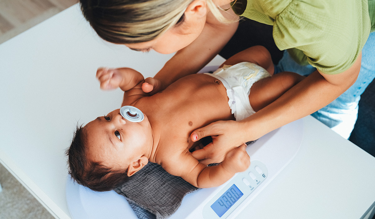 A mother carefully weighs her baby on a digital scale at home, monitoring the infant’s growth and development. The baby lies comfortably in a diaper while the mother gently supports and reassures them. The scene highlights attentive parental care, regular health monitoring, and the importance of tracking a baby’s weight in a nurturing home environment.