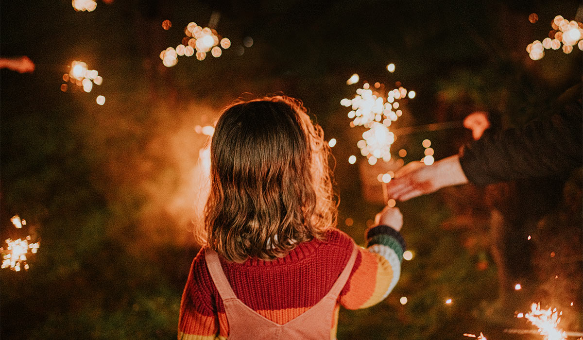 A young child holds a burning sparkler in the evening. View is from the back of her head as she holds the handheld firework. Darkness provides a space for copy.