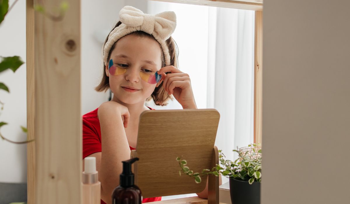 A teenage girl gently cares for her skin, placing soothing patches under her eyes as she applies facial products at home, all while gazing into the mirror