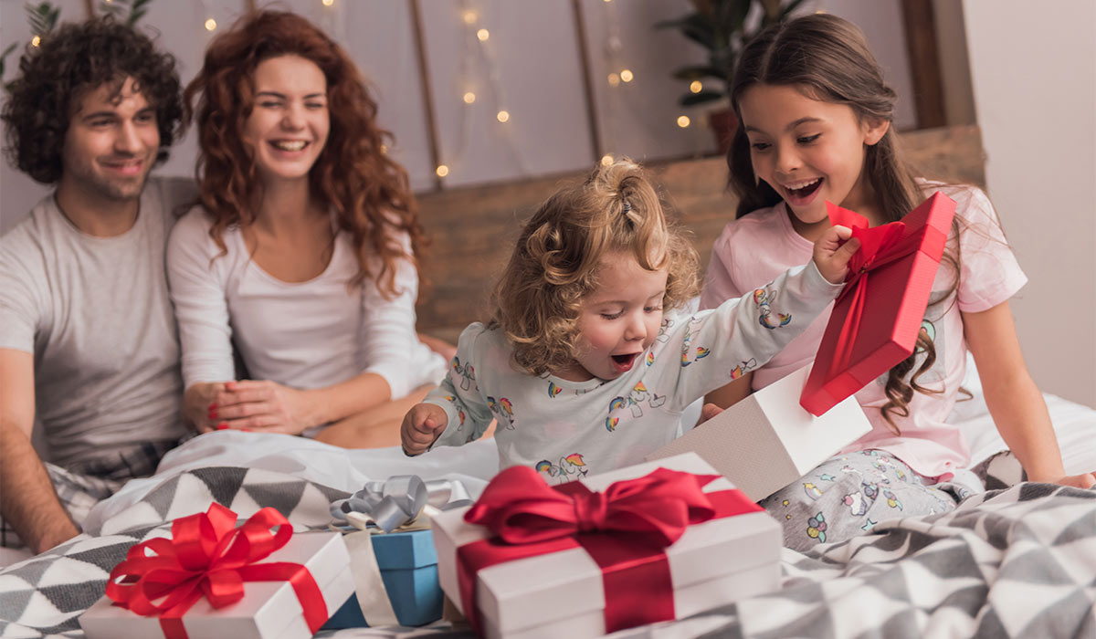 It's Christmas time! Happy family sitting in bed, parents are smiling while their daughters are opening the presents; Shutterstock ID 777730984; purchase_order: -; job: -; client: -; other: