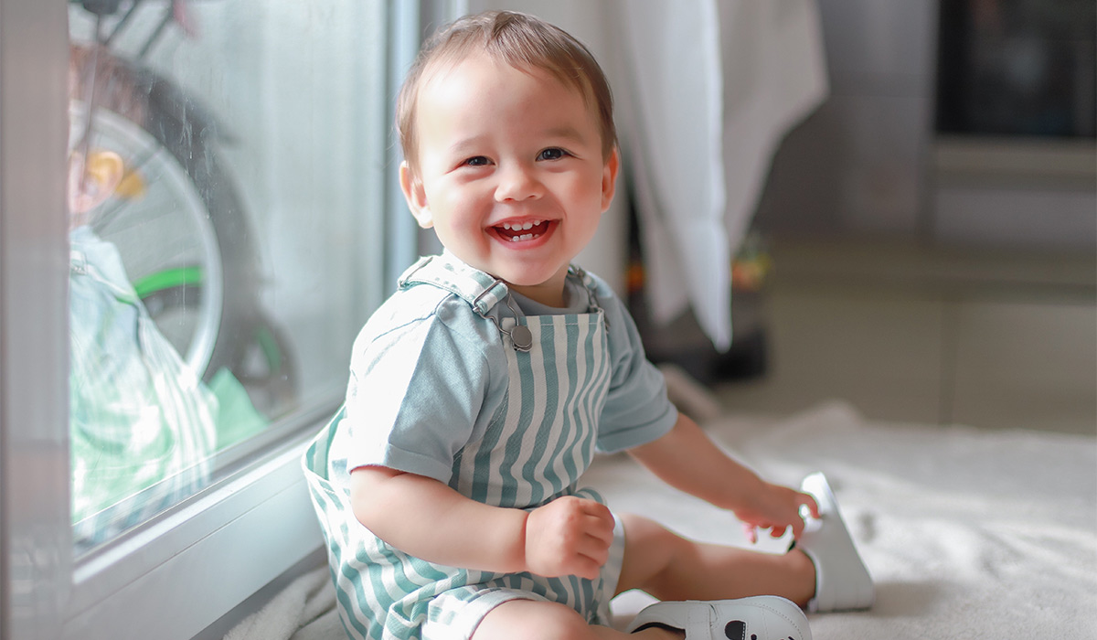 Happy laughing baby boy with teething wearing green dress sitting next to window home.Portrait of mixed race Asian-German infant about one year old joyful with copy space for text.; Shutterstock ID 1751913986; purchase_order: -; job: -; client: -; other: