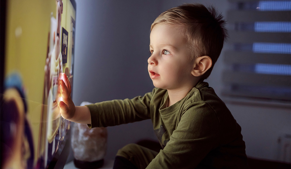Children's addiction to television and cartoons. A boy touches the TV screen with one hand. Close up of a kid sitting right in front of the TV in his pajamas and staring at a cartoon. Modern parenting; Shutterstock ID 1968307801; purchase_order: -; job: -; client: -; other: