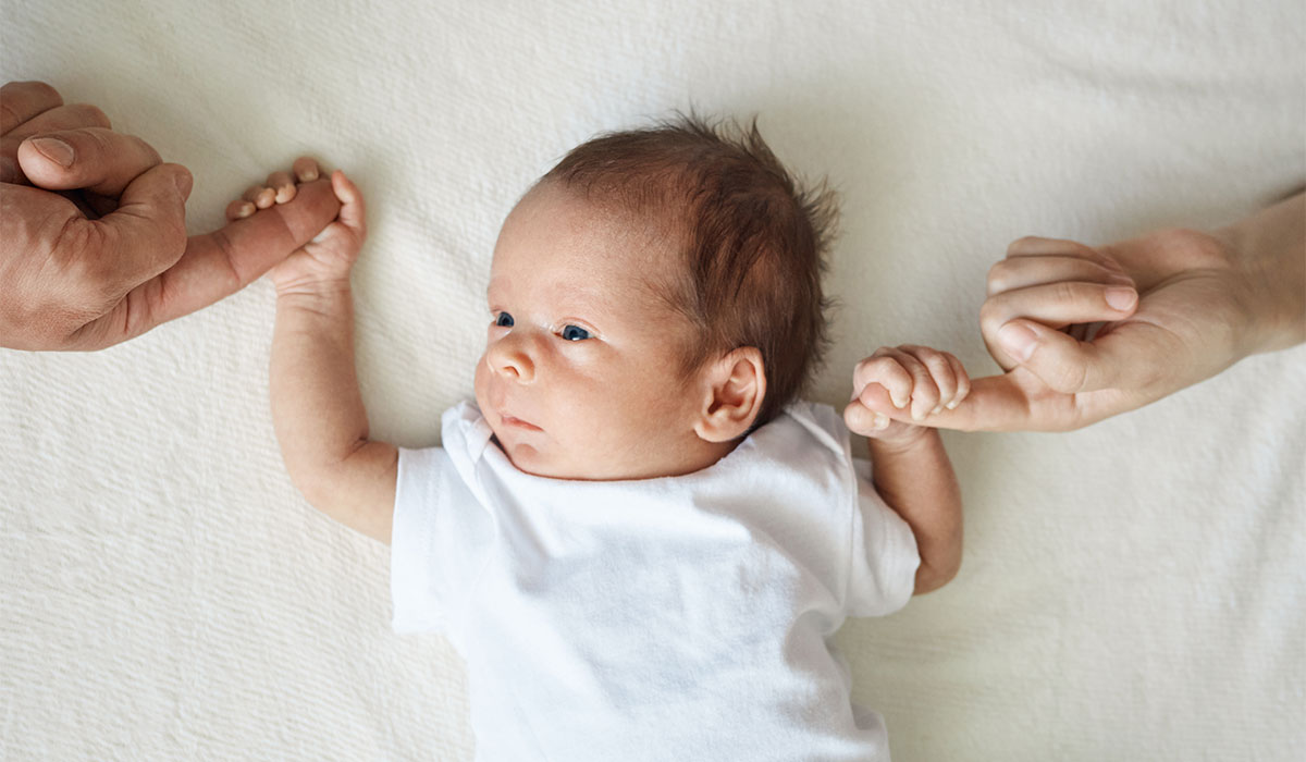 Baby holding parents hands. Father and mother hold newborn kid cute infant child in white bodysuit lying on white sheet on bed; Shutterstock ID 2475571229; purchase_order: -; job: -; client: -; other: