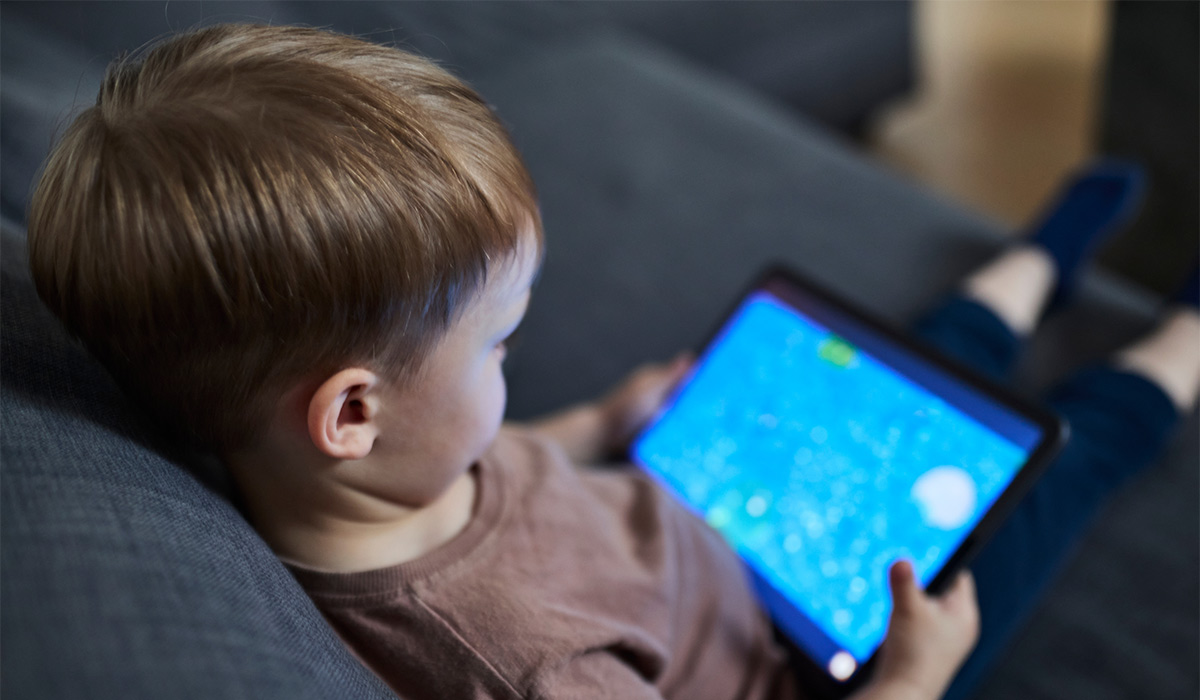 Curious three-year-old boy sitting comfortably on a sofa while watching a tablet screen. Focused expression on his face as he engages with digital content. The child is dressed in casual clothes,