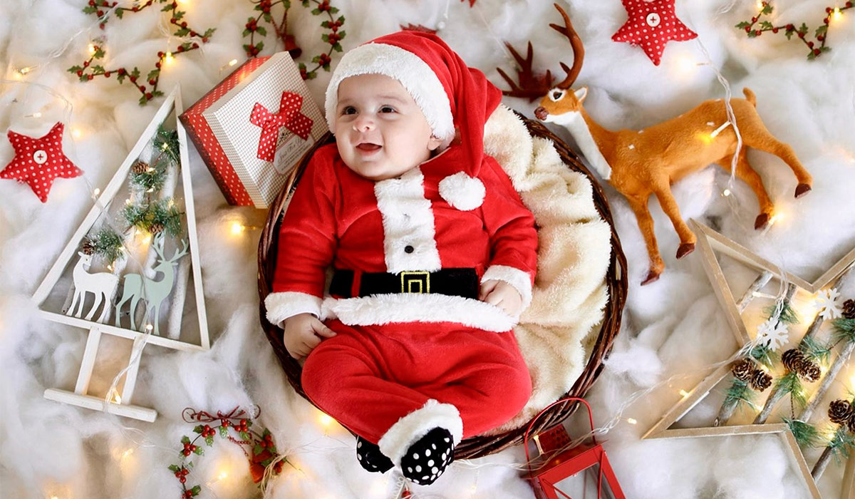 Adorable baby in Santa Claus costume surrounded by Christmas decorations and lights.