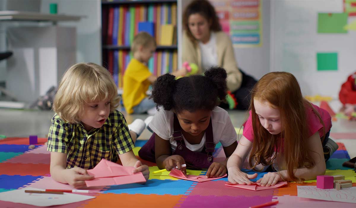 Little kids learn to folding paper origami lying on floor in kindergarten. Diverse preschool children doing craft at nursery school playroom; Shutterstock ID 2319071907; purchase_order: -; job: -; client: -; other: