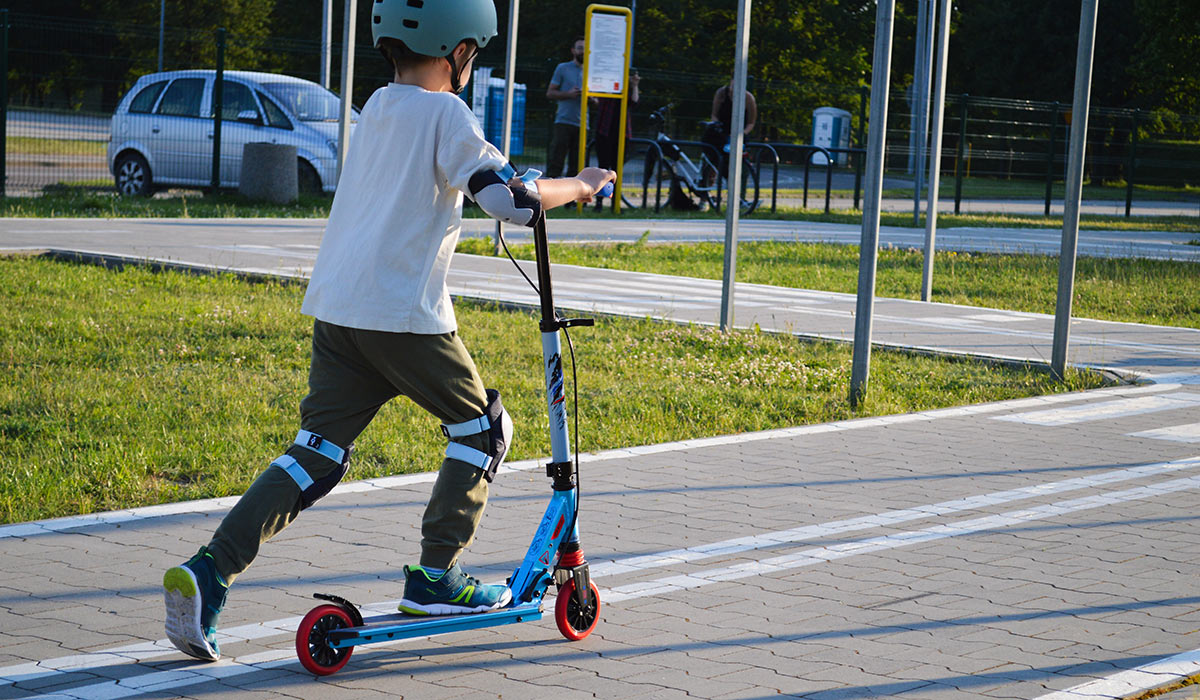 Image of a child wearing knee and elbow pads riding a blue scooter on a sidewalk near a road with cars parked. Photo emphasizes child safety while playing near traffic, importance of protective gear; Shutterstock ID 2685220923; purchase_order: -; job: -; client: -; other: