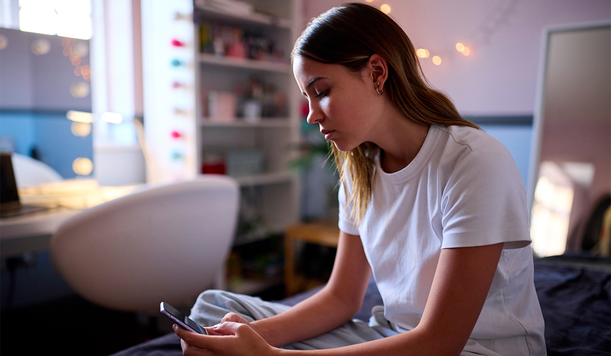 Depressed Teenage Girl Sitting On Bed At Home Looking At Mobile Phone; Shutterstock ID 2261420981; purchase_order: -; job: -; client: -; other: