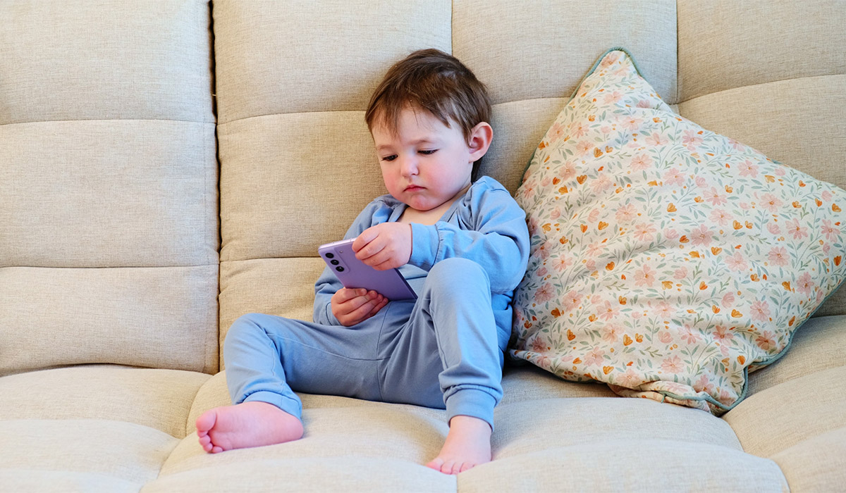 Toddler baby is tapping away on the mobile phone screen. The happy youngster is glued to the screen of their phone while seated comfortably on the couch. Kid aged about two years (one year ten months); Shutterstock ID 2307643563; purchase_order: -; job: -; client: -; other: