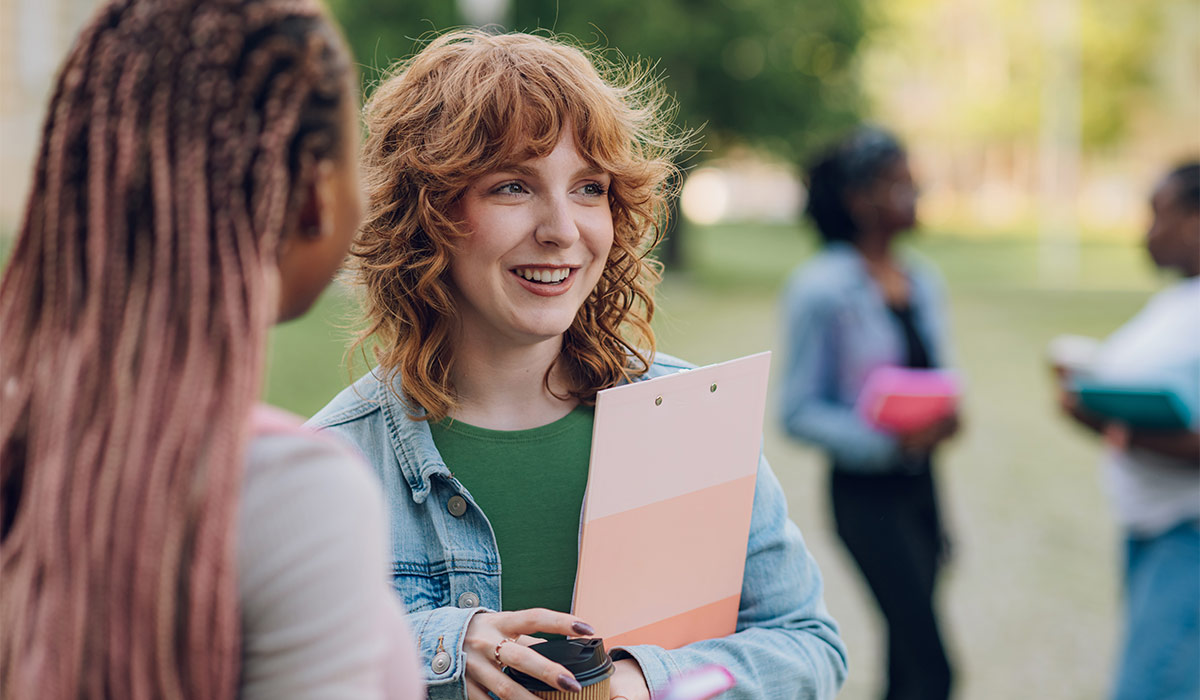 Portrait of smiling college student standing at university campus with her multicultural female classmate with clipboard and coffee to go in hands and discussing school project. Students on a break.; Shutterstock ID 2474168695; purchase_order: -; job: -; client: -; other: