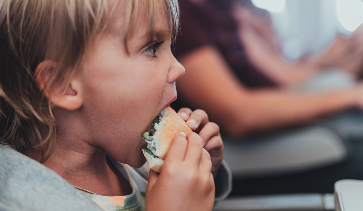 little candid kid boy five years old eats burger or sandwich food sitting in airplane seat on flight traveling from airport. children take a bite. child in air plane eating lunch or dinner meal; Shutterstock ID 2203196615; purchase_order: -; job: -; client: -; other:
