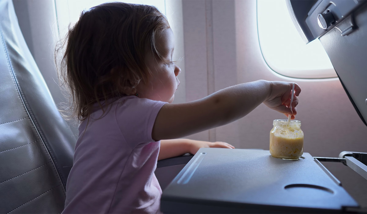 Preschooler girl eats fruit puree with spoon during airplane flight. Cute little child licks spoon with pleasure enjoying tasty snack; Shutterstock ID 2577617975; purchase_order: -; job: -; client: -; other: