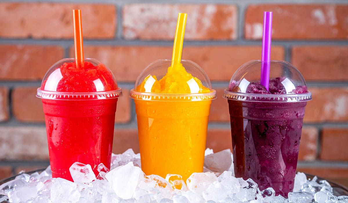 Three vibrant frozen fruit slushies in plastic cups with straws, nestled on ice against a brick backdrop. Red, orange, and purple colors are shown.; Shutterstock ID 2623766955; purchase_order: -; job: -; client: -; other: