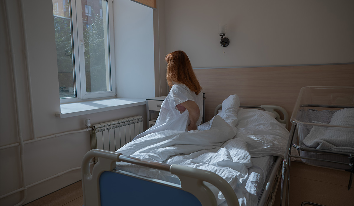 An unhappy woman sits on a couch and holds her stomach in a hospital room after giving birth.; Shutterstock ID 2596681763; purchase_order: -; job: -; client: -; other: