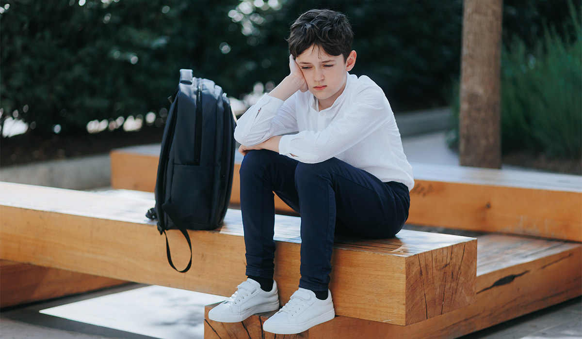 Tired and sad school-aged boy in uniform sits quietly in a schoolyard during break. His expression shows emotional distress, possibly from bullying or school pressure.; Shutterstock ID 2647926361; purchase_order: -; job: -; client: -; other:
