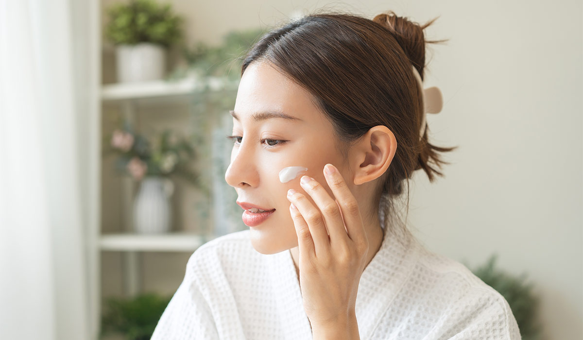Facial beauty skin care, smile of pretty asian young woman in bathrobe looking at mirror, hand applying moisturizer lotion on her face, putting cream treatment before makeup cosmetic routine at home.; Shutterstock ID 2684948347; purchase_order: -; job: -; client: -; other: