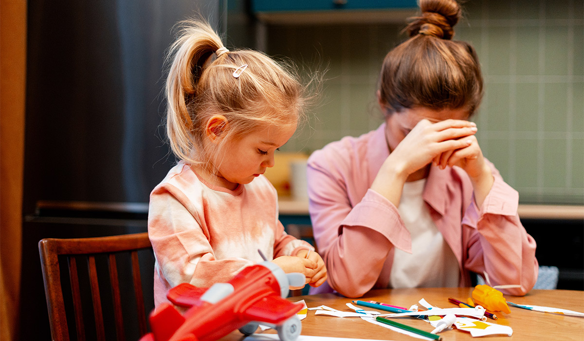 Stressed mother covering face with hands while little girl playing with toy airplane at kitchen table; Shutterstock ID 2717476999; purchase_order: -; job: -; client: -; other: